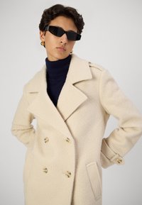 Krēms krāsas divrindu mētelis, izgatavots no teksturēta auduma, ar lieliem uzlāgiem, zelta pogām un sānu kabatām. Valkāts virs tumši zilā turtleneck.