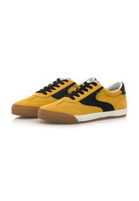 Zapatillas amarillas con acentos en negro y blanco, que presentan una tela texturizada, punta redonda y un diseño con cordones, apoyadas sobre una suela de goma.
