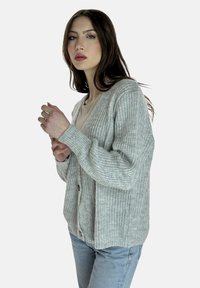 Cardigan a maglia grigio chiaro con scollo a V, chiusura con bottoni e trama a coste; abbinato a una camicia crema e jeans azzurri.