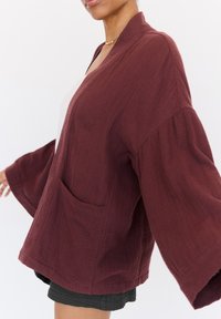 Veste oversize bordeaux en tissu texturé avec des manches larges et une poche avant, présentant un design décontracté et à ouverture devant.