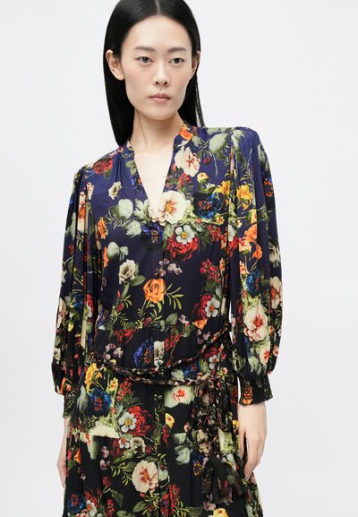 Lágyan áramló, virágmintás ruha mély V-nyakkivágással, hosszú ballonujjakkal és derékban megkötve. Sötét háttér, élénk színű, sokszínű virágokkal.