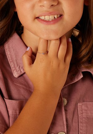 Zilveren ring met een kleine blauwe edelsteen, gedragen aan de hand van een kind, tegen een achtergrond van een roze stoffen shirt. Glad oppervlak en eenvoudig ontwerp.