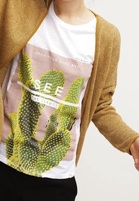 Un long cardigan couleur moutarde porté sur un t-shirt blanc avec un motif de cactus en vert et jaune, accompagné d'un texte imprimé en noir et d'accents rose.