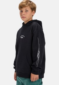 Chlapec so svetlo hnedými vlasmi, oblečený v čiernej mikine Quiksilver s bielym dizajnom na rukávoch a tyrkysovými nohavicami, stojí pred jednoduchým pozadím.