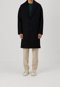 Schwarzer Wollmantel mit gekerbtem Reverskragen, zwei Knöpfen und geradem Schnitt. Kombiniert mit beigen Hosen und weißen Turnschuhen.