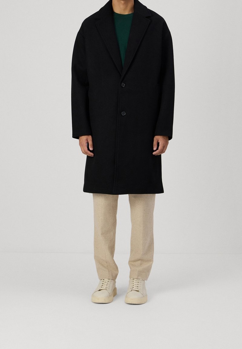 Schwarzer Wollmantel mit gekerbtem Reverskragen, zwei Knöpfen und geradem Schnitt. Kombiniert mit beigen Hosen und weißen Turnschuhen.