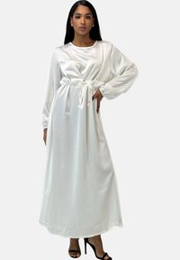 Witte satijnen maxi-jurk met lange mouwen, taillekoord en losse pasvorm. Glad oppervlak en vloeiend ontwerp. Zwarte sandalen met hak.