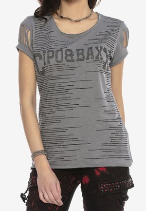 Kvinde iført en grå T-shirt med sort prikket tekst "TIPO & BAXX" og udskæringsdetaljer på ærmerne, kombineret med sorte og røde nittebukser.