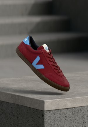 Piros bőr sneaker kék részletekkel, barna talppal és fűzőkkel, betonfelület fölött lebegve, háttérben elmosódott lépcsőkkel.