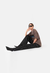 Przesadna brązowa koszulka z niebieskimi grafikami, połączona z czarnymi spodniami cargo. Modelka siedzi w eleganckich czarnych okularach przeciwsłonecznych i z warstwową biżuterią.