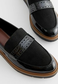 Mocassini neri con tomaia in suede, dettagli in vernice lucida, doppie cinghie texturizzate con ritagli circolari e suola in gomma resistente.