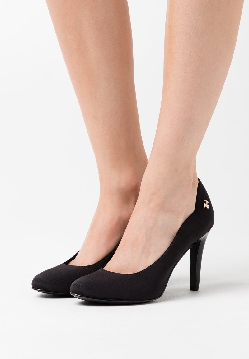 Escarpins noirs à talons hauts, en tissu lisse, avec un embout arrondi et des bords légèrement festonnés. Un petit logo métallique est présent sur le talon.