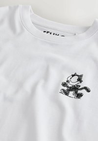 T-shirt bianca a girocollo con un piccolo schizzo di un gatto nero in stile cartone animato sul petto, con grandi occhi e una zampa sollevata.