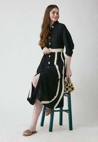 Schwarzes Leinenkleid mit weißen Streifenakzenten, übergroßen Knöpfen und einem Gürtel. Model sitzt auf einem grünen Stuhl und hält eine mit Perlen besetzte goldene Handtasche.
