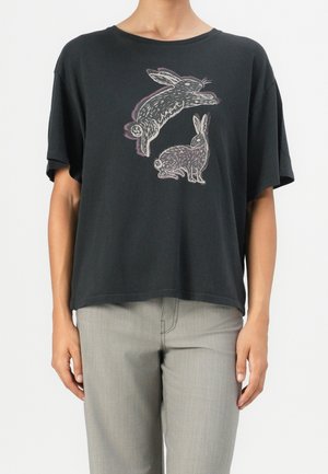 Czarny, luźny T-shirt z dwoma ilustrowanymi królikami, jednym skaczącym i drugim siedzącym, zestawiony z jasnoszarymi spodniami.