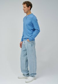 Maglietta henley a maniche lunghe di colore blu chiaro con colletto rotondo, abbinata a jeans blu chiaro a vestibilità ampia e sneakers bianche, che mostrano uno stile casual.