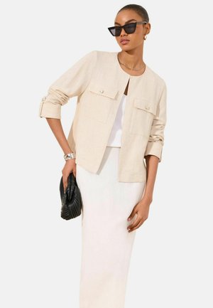 Nő bézs gombos zakót visel fehér ruha fölött, fekete macskaszemu napszemüveg, arany karika fülbevaló, ezüst karkötő, fekete kis táskát tart.