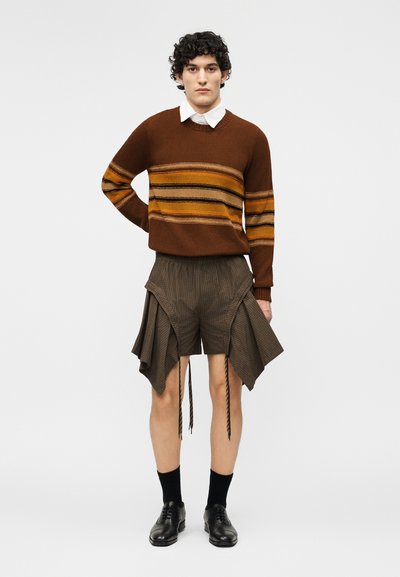 Jeune homme aux cheveux bouclés portant un pull rayé marron, une chemise blanche à col, un short marron asymétrique, des chaussettes noires et des chaussures noires.