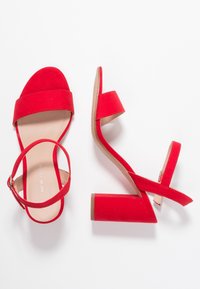 Red suede heeled sandals with a wide toe strap, adjustable ankle strap, and block heel. Smooth inner lining and textured sole.