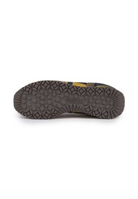 Suela de zapato de goma marrón con patrón de agarre texturizado, que presenta formas circulares y de diamante, y un detalle de logo central. Acentos en amarillo y azul marino visibles.
