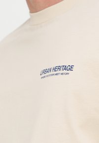 T-shirt beige avec un texte bleu sur la poitrine indiquant "URBAN HERITAGE WHERE FOOTSTEPS MEET HISTORY", porté par une personne.