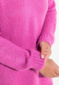 Pull en tricot rose avec une coupe ample, un matériau doux et texturé, et des poignets côtelés. Gros plan sur le détail de la manche et une main tenant l'ourlet.