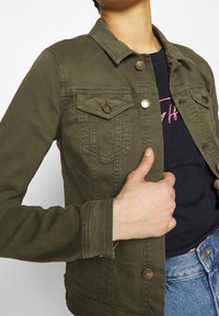 Olivgrüne Denimjacke mit zwei Brusttaschen, bronzefarbenen Knöpfen und kontrastierender Naht, offen getragen über einem schwarzen Hemd und blauen Jeans.