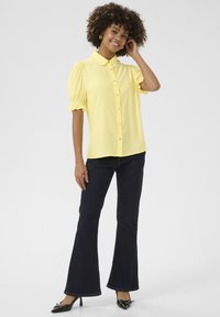 Blusa gialla con colletto e maniche a palloncino, chiusura a bottoni, abbinata a jeans neri a zampa d'elefante e tacchi neri. Tessuto semplice e liscio.