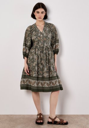 Vestido verde con estampado de paisley, con escote en V, mangas abullonadas y cintura fruncida. Presenta un dobladillo estampado y se combina con sandalias marrones.