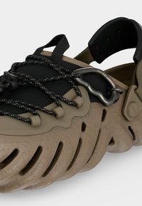 Μπεζ και μαύρο παπούτσι Croslite με ανοιχτές πλευρές, ρυθμιζόμενα μαύρα κορδόνια και καραμπίνερ με την ένδειξη "Όχι για αναρρίχηση" στο πάνω μέρος.