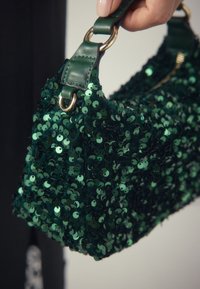 Sac à main vert à paillettes avec une surface texturée, doté d'une poignée en cuir et d'accents en métal doré, présentant une forme compacte et rectangulaire.