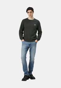 Felpa verde scuro con colletto bianco a contrasto, abbinata a jeans azzurri e scarpe nere. Modello in piedi con le mani in tasca.