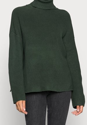 Pull à col roulé en maille côtelée vert foncé porté avec un pantalon noir, manches longues et coupe légèrement ample.