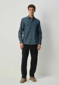 Košeľa na gombíky v tmavomodrej farbe s geometrickým vzorom, v kombinácii s čiernymi nohavicami a svetlo hnedými topánkami, so voľným strihom a dlhými rukávmi.