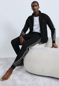 Čierna tepláková súprava z mäkkého materiálu s bielymi a sivými akcentmi, bunda so zipsom a zúžené nohavice. Model sedí na textúrovanom povrchu.