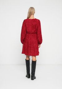 Frau mit langen blonden Haaren, die ein strukturiertes rotes knielanges Kleid und schwarze kniehohe Stiefel mit Absatz trägt, mit dem Rücken zu einer weißen Hintergrund.