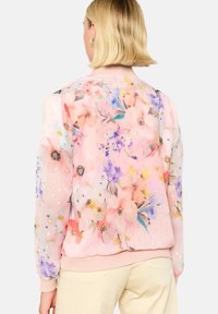 Blommig bomberjacka i genomskinlig rosa tyg, med färgglada blommor och subtila metalliska detaljer. Ribbad mudd och nederkant. Lätt design.