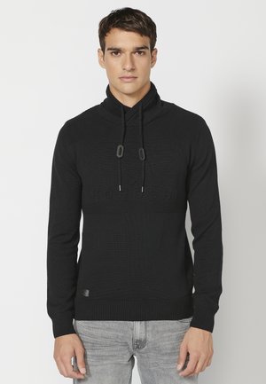 Jersey de punto negro con cuello alto y cordones. Presenta una sección superior texturizada y un detalle del logo en la parte frontal. Puños y dobladillo acanalados.