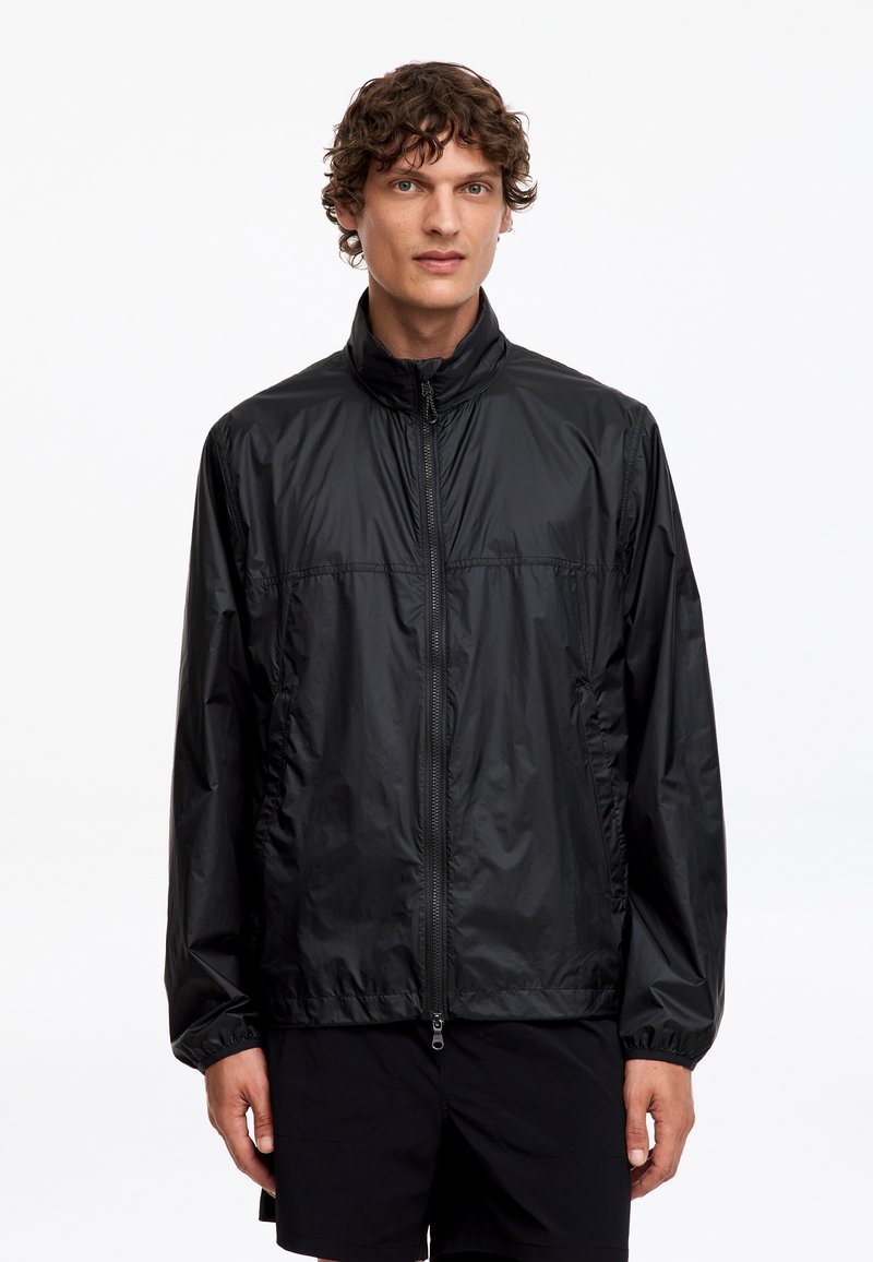 Homme aux cheveux bouclés porte une veste zippée noire légère et un short noir, debout devant un fond blanc uni.