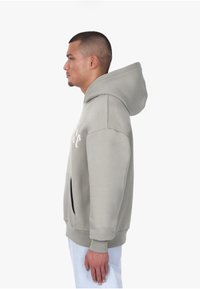 Sweat à capuche gris clair avec poche avant, poignets côtelés et grande capuche. Présente un logo blanc sur la poitrine. Texture de tissu doux.