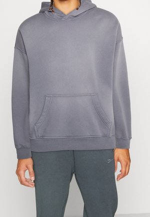 Person trägt ein schlichtes graues Kapuzensweatshirt mit Kängurutasche und dunkelgraue Hose, steht vor einem hellen Hintergrund.