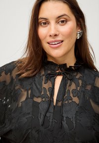 Sort gennemsigtig bluse med blomsterbroderi, flæset krave og bindebånd. Bemærkelsesværdige teksturkontraster med baggrunden og blomsterdesignene.