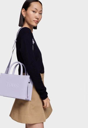Bolso tote de cuero lila con logo "TOUS" en relieve, dos asas superiores y correa de hombro desmontable. Estilizado con un suéter negro y una falda beige.