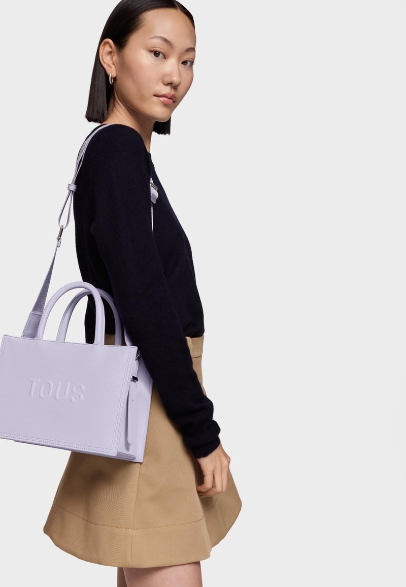 Borsa tote in pelle lilla con logo "TOUS" inciso, doppio manico superiore e tracolla removibile. Abbinata a un maglione nero e una gonna beige.