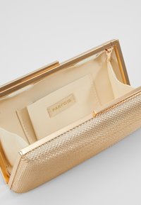 Clutch dorato metallizzato con superficie testurizzata, dotato di una fodera interna liscia e un'etichetta del marchio. Include una chiusura a cornice.