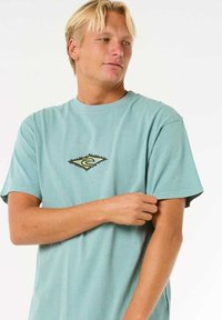 Himmelblaues T-Shirt mit kurzen Ärmeln aus Baumwolle. Mit einem kleinen, gelb-schwarzen Logo in Diamantform auf der Brust.