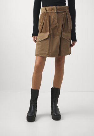 Femme portant une jupe trapèze en velours côtelé et tissu beige avec de grandes poches avant, une ceinture, un top noir et des bottines plateformes noires.