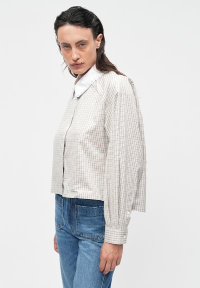 Cropped button-up shirt in light beige with a check pattern, featuring a white collar and billowy long sleeves. Paired with blue jeans.