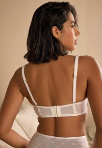 Reggiseno in rete bianca con spalline regolabili e finiture in pizzo. Presenta una chiusura con gancio e occhiello. Lo sfondo è di un morbido beige.