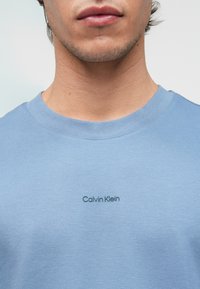 Világoskék pamut póló kerek nyakkal; a mellkas bal oldalán egy kisebb, sötétszürke "Calvin Klein" logó található.